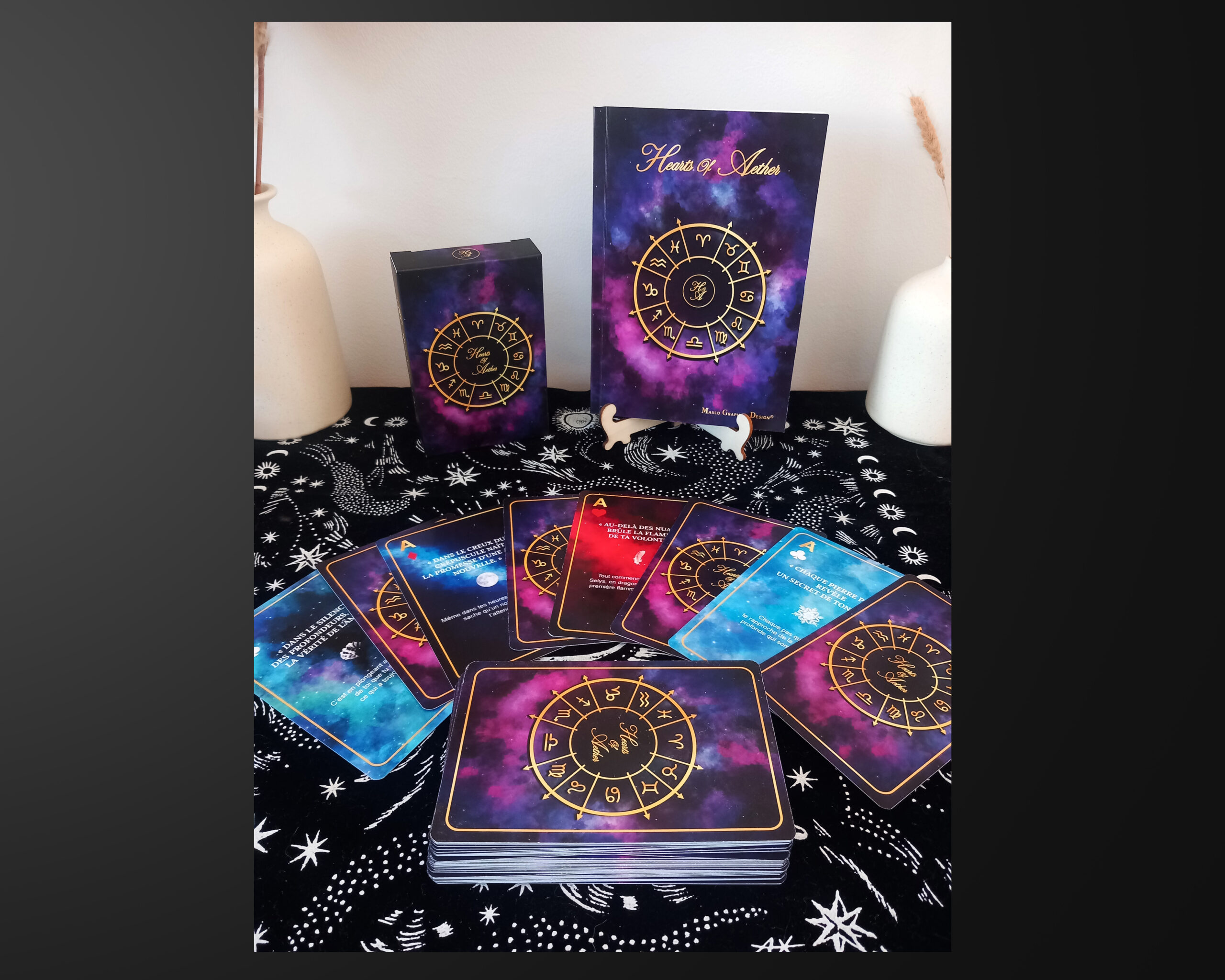 Photographie d’un coffret de cartes inspiré de l’astrologie, composé d’une boîte, d’un livret illustré et de cartes aux motifs cosmiques violets, bleus et dorés disposées sur un tissu céleste.