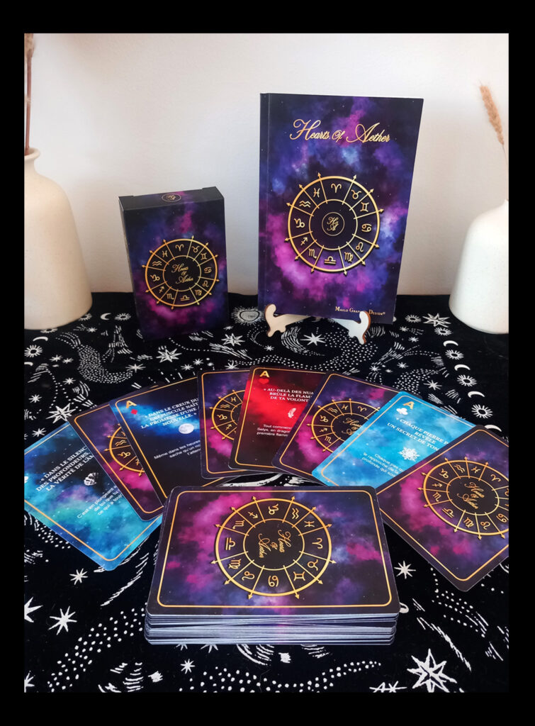 Photographie d’un coffret de cartes inspiré de l’astrologie, composé d’une boîte, d’un livret illustré et de cartes aux motifs cosmiques violets, bleus et dorés disposées sur un tissu céleste.